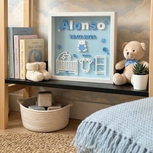 Könnte beinhalten: Ein Baby-Ankündigungsrahmen mit dem Namen "Alonso" in Blau und dem Datum 28.02.2026. Der Rahmen enthält einen Teddybären, ein Kinderbett, einen Strampler und andere Baby-Elemente. Ein Teddybär und eine kleine Topfpflanze stehen auf dem Regal.