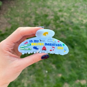 May include: Pale blue hair claw clip with the text "In my Boston Marathon Era" and various icons. The clip features a unicorn, a shoe, a musical note, and a map of Massachusetts. The text "Hereford St Boylston St" is also visible.