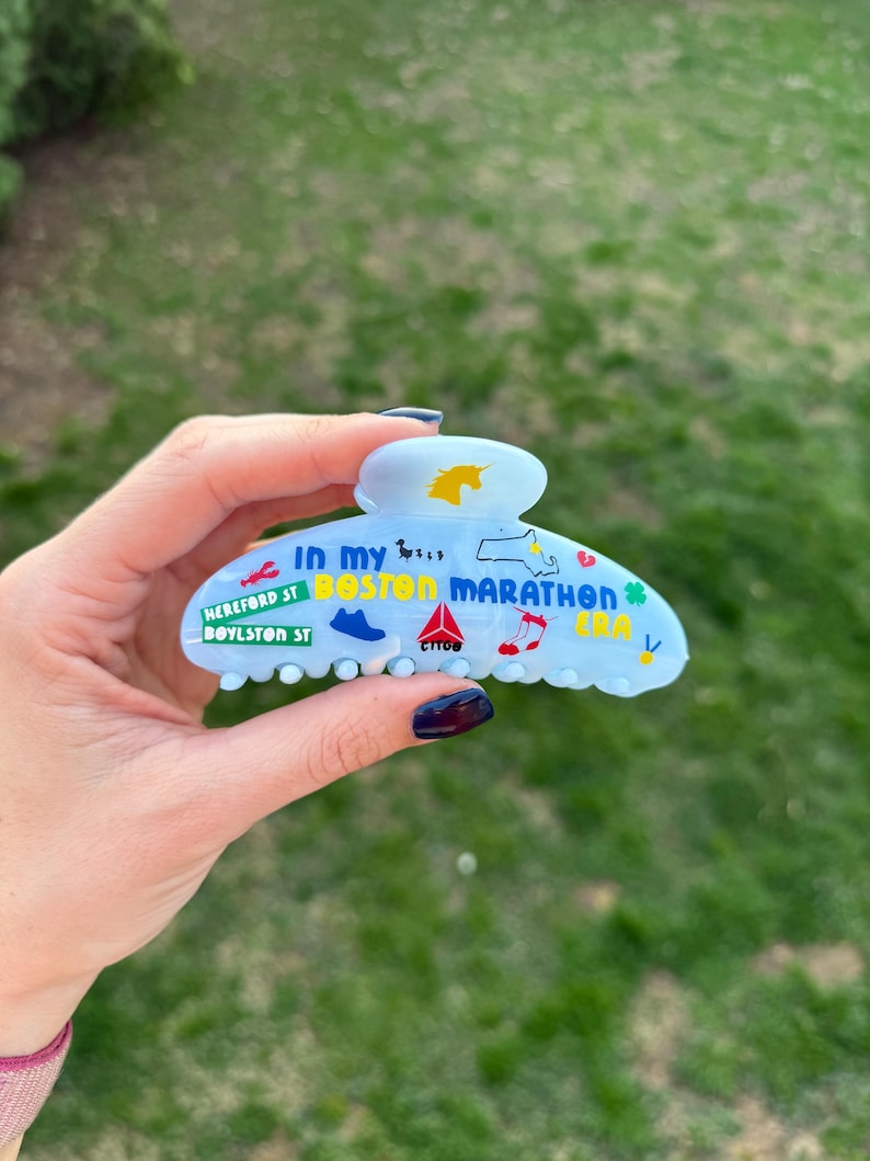 May include: Pale blue hair claw clip with the text "In my Boston Marathon Era" and various icons. The clip features a unicorn, a shoe, a musical note, and a map of Massachusetts. The text "Hereford St Boylston St" is also visible.