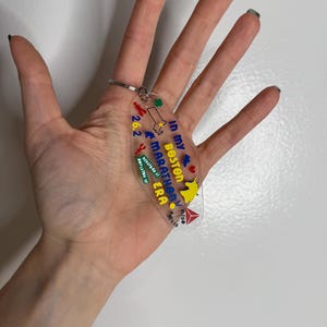 May include: Clear acrylic keychain with a silver keyring. The keychain features colorful text and graphics, including the words "In my Boston Marathon Era" and "26.2". The design includes a map of Massachusetts and other marathon-related icons.