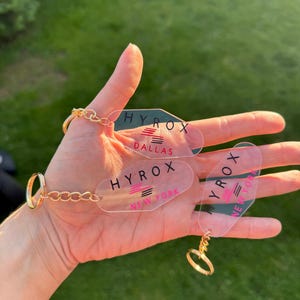 May include: Transparent acrylic keyrings with gold-coloured rings and chains. Each keyring features the word "HYROX" in black, with pink stripes and the city names Dallas and New York.