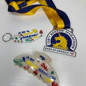 May include: A selection of Boston Marathon souvenirs, including a medal with a blue and yellow ribbon, a keyring, and a hair clip. The medal displays a unicorn and the text "128th Boston Marathon 2024". The keyring and hair clip have the text "In My Boston Marathon ERA".