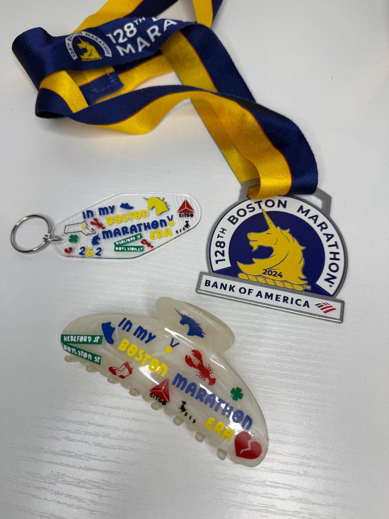 May include: A selection of Boston Marathon souvenirs, including a medal with a blue and yellow ribbon, a keyring, and a hair clip. The medal displays a unicorn and the text "128th Boston Marathon 2024". The keyring and hair clip have the text "In My Boston Marathon ERA".