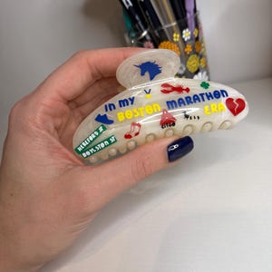 May include: A cream-coloured hair claw clip with colourful graphics and text. The clip features a blue unicorn, a red lobster, and the words "IN MY BOSTON MARATHON ERA." Additional text includes "HEREFORD ST BOYLSTON ST" and "CITGO."