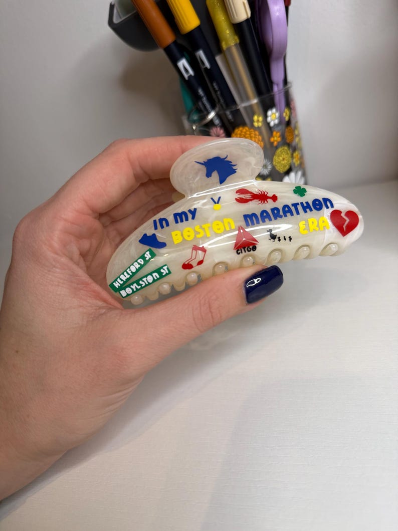 May include: A cream-coloured hair claw clip with colourful graphics and text. The clip features a blue unicorn, a red lobster, and the words "IN MY BOSTON MARATHON ERA." Additional text includes "HEREFORD ST BOYLSTON ST" and "CITGO."