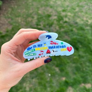 May include: A pale blue hair claw clip decorated with colourful graphics and text, including a unicorn, lobster, and the words "Boston Marathon." The clip also features the text "Hereford St, Boylston St."
