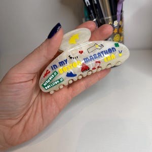 May include: White marbled hair claw clip with colourful Boston Marathon-themed graphics and text. The clip features illustrations of a unicorn, a map of Massachusetts, and the text "IN MY BOSTON MARATHON ERA".