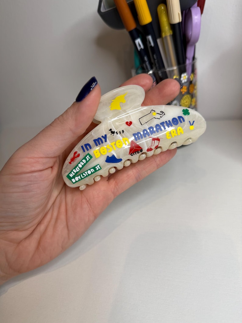 May include: White marbled hair claw clip with colourful Boston Marathon-themed graphics and text. The clip features illustrations of a unicorn, a map of Massachusetts, and the text "IN MY BOSTON MARATHON ERA".
