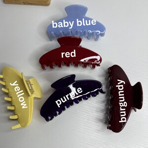 May include: Six claw hair clips in baby blue, red, yellow, purple, burgundy, and a darker burgundy. Each clip is labelled with its colour. The clips are displayed on a white surface.