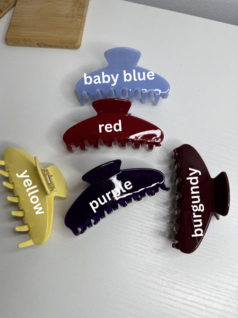 May include: Six claw hair clips in baby blue, red, yellow, purple, burgundy, and a darker burgundy. Each clip is labelled with its colour. The clips are displayed on a white surface.