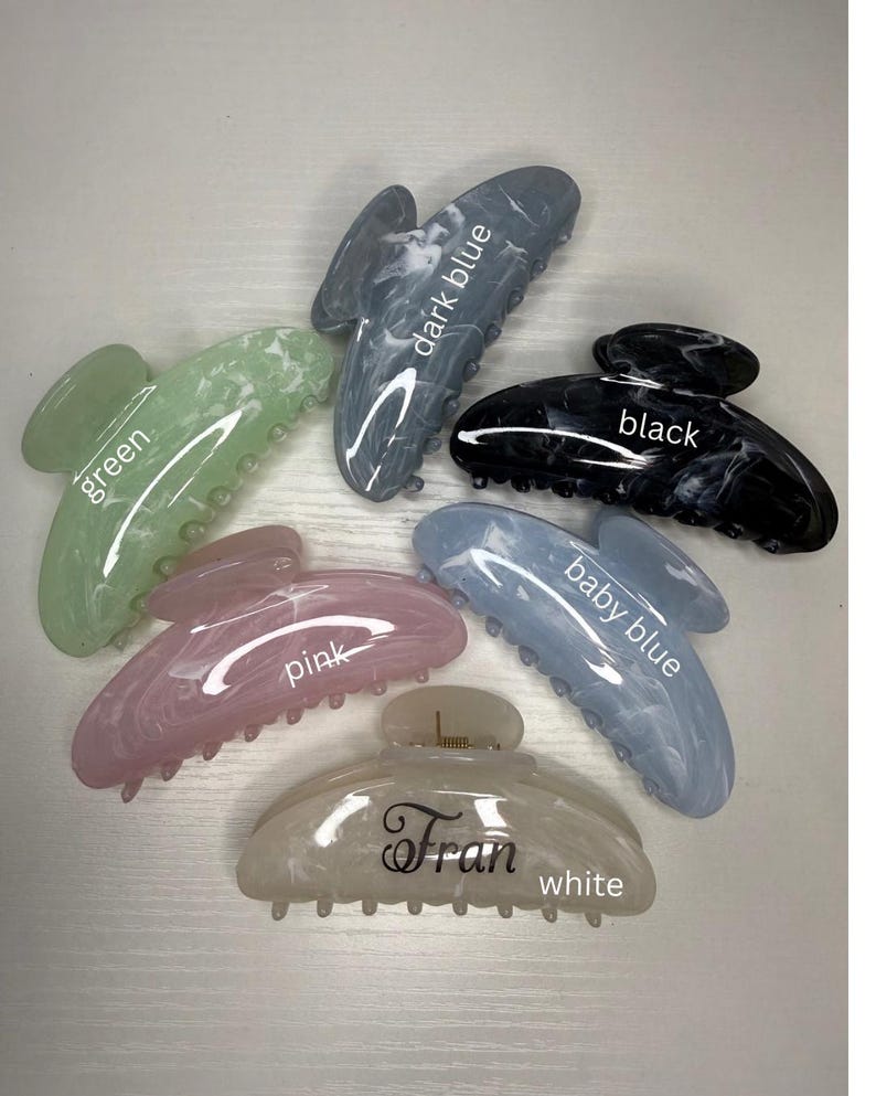 May include: Six marbled hair claw clips in shades of green, pink, baby blue, dark blue, black, and white. Each clip is labelled with its colour. The white clip has the name "Fran" printed on it.