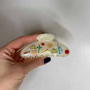 Op de afbeelding: Crème-witte haarclip met New York City-thema ontwerpen. De clip heeft illustraties van een gele taxi, een appel, een pretzel, het Vrijheidsbeeld en de tekst "I NY".
