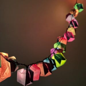 Puede incluir: Una cadena de faroles de papel origami de colores, rojos, verdes, rosas y amarillos. Las formas geométricas están iluminadas desde dentro, creando un brillo ambiental cálido. Los faroles están ensartados en un cable negro, perfectos para la decoración festiva.