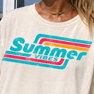 Peut inclure: T-shirt crème avec l'inscription "Summer Vibes" en turquoise, orange et rouge dans une police rétro. Le t-shirt a des manches courtes et une coupe décontractée. Un van vintage bleu clair et blanc est en arrière-plan.