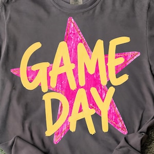 Op de afbeelding: Grijs t-shirt met een roze ster en de tekst "GAME DAY" in het geel. Het shirt heeft een ronde hals en is gemaakt van zacht materiaal.