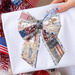Könnte beinhalten: Ein weißes T-Shirt mit einer großen patriotischen Schleife. Die Schleife hat ein Patchwork-Design mit amerikanischer Flagge und historischen Bildern. Das Bild enthält rot-weiß-blaue Dekorationen, Miniaturflaggen und eine Discokugel.