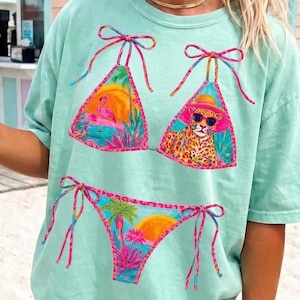 May include: A light green t-shirt featuring a colorful bikini design. The bikini top and bottom are adorned with tropical scenes, including a flamingo, leopard, and palm trees. The shirt has a relaxed fit and is suitable for casual wear.