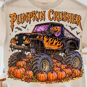 Könnte beinhalten: Ein cremefarbenes T-Shirt mit einer Grafik eines schwarzen Monstertrucks mit orangefarbenen Flammen, gefahren von einer Figur mit Kürbiskopf. Der Text PUMPKIN CRUSHER steht über dem Truck, umgeben von orangefarbenen Kürbissen und Herbstlaub.