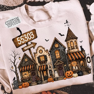 May include: A cream-colored crewneck sweatshirt featuring a graphic of a spooky Halloween village with pumpkins, ghosts, and a signpost labeled 55303 Minnesota. The design includes a witch on a weather vane, bats, and a bare tree against a dark sky.