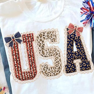Könnte beinhalten: Weißes T-Shirt mit dem Wort "USA" in rot-weiß gestreiften, Leopardenmuster und marineblauen Applikationen, mit Spitzenbesatz. Die Buchstaben sind mit Schleifen verziert. Patriotisches Thema.