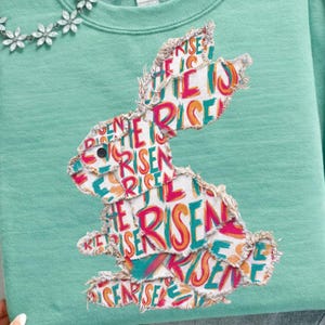 May include: A mint green sweatshirt with a distressed bunny graphic, featuring the words "He Is Risen". The design is in a patchwork style. A silver floral necklace and a brown hat are also visible.