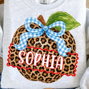 Peut inclure: Un sweat-shirt gris clair à col rond orné d'un motif central représentant une citrouille au pelage léopard marron et noir. La citrouille est décorée d'un nœud vichy bleu et blanc et d'une feuille verte, avec le nom SOPHIA en lettres blanches.