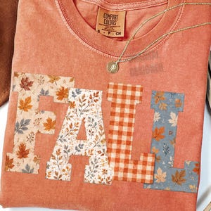 May include: A terracotta-colored short-sleeve t-shirt with a crew neckline featuring the word FALL in large block letters across the chest. Each letter has a distinct pattern: autumn leaves, floral foliage, orange gingham, and blue leaves.