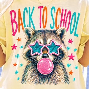 May include: A pale yellow t-shirt featuring a graphic of a raccoon wearing star-shaped sunglasses and blowing a pink bubble. The text "BACK TO SCHOOL" is printed in colourful letters above the raccoon, surrounded by small orange and pink stars.