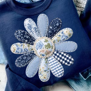 Puede incluir: Una sudadera azul marino con una gran margarita aplicada. Los pétalos de la margarita están hechos de varias telas azules estampadas, incluyendo diseños florales, a rayas y de cuadros. El centro de la flor es un estampado floral con un ribete de encaje color crema.