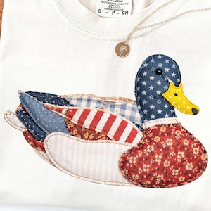 Puede incluir: Camiseta color crema con un diseño de pato patchwork con patrones patrióticos. El pato tiene una cabeza azul con estrellas blancas, un pico amarillo y alas y cuerpo en telas rojas, blancas y azules. Un collar dorado con un colgante descansa sobre la camisa.