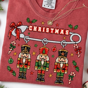 May include: A brick red t-shirt featuring a large safety pin graphic with the word CHRISTMAS in red blocks. Three nutcracker figures in red and green uniforms hang from the pin, surrounded by small stars, holly, and candy cane illustrations.
