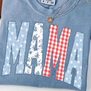 May include: A blue short-sleeve t-shirt with a crew neckline and a Comfort Colors label. The front features the word MAMA in large block letters with raw edges. The letters have patterns: blue with white stars, white with blue florals, and red gingham.