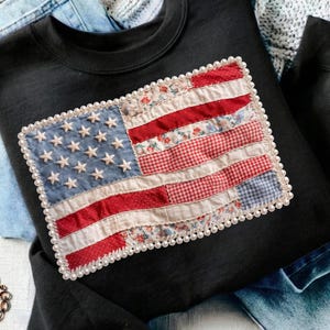 Könnte beinhalten: Schwarzes Sweatshirt mit einem Patchwork-Design der amerikanischen Flagge. Die Flagge besteht aus verschiedenen gemusterten Stoffen, darunter Blumenmuster und Vichy, und hat eine perlenartige Verzierung. Die Sterne der Flagge sind weiß, die Streifen rot und weiß.
