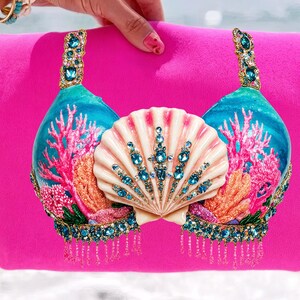 May include: A hot pink bikini top with a seashell design. The turquoise cups feature coral and are embellished with blue gems. A large, white seashell is centered between the cups. The straps and bottom edges are also adorned with gems.