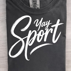 May include: A charcoal gray crew neck t-shirt featuring the white text "Yay Sport" in a stylized, brush-stroke script font across the front. The garment includes a visible interior neck label with the brand name Comfort Colors printed on it.