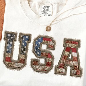 Op de afbeelding: Crèmekleurig T-shirt met het woord "USA" in een patchwork-ontwerp met de Amerikaanse vlag en luipaardprint. Het shirt heeft een ronde hals en een label met "Comfort Colors". Een gouden ketting met een hanger is over het shirt gedrapeerd.