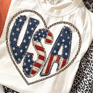 May include: An off-white t-shirt featuring a large heart graphic outlined in a leopard print pattern. Inside the heart, the letters USA are displayed in a distressed, frayed-edge fabric design with a blue star and red and white stripe motif.
