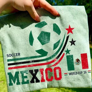 May include: A light green t-shirt featuring a graphic of a soccer ball, the text "SOCCER MEXICO" in green, black, and red, a small Mexican flag, and the text "WORLD CUP '26" with three stars in red, black, and green above the flag.