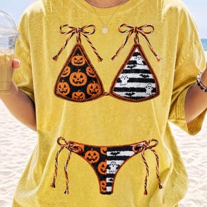 May include: Yellow t-shirt with a Halloween bikini design. The bikini top features pumpkin and ghost patterns, with matching bottoms. The design is in black and orange, ideal for the spooky season.