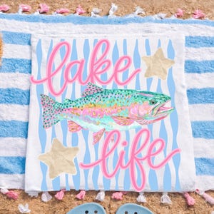 May include: White beach towel with a colorful fish illustration, the words "Lake Life" in pink script, and two beige starfish. The towel has blue and white stripes and is on a sandy beach.