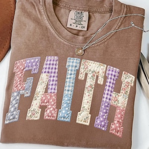 Könnte beinhalten: Ein braunes Comfort Colors T-Shirt mit dem Wort "FAITH" in einem Patchwork-Design appliziert. Die Patchwork-Buchstaben weisen Blumen-, Vichy- und Tupfenmuster in Pastellfarben auf. Eine silberne Halskette mit einem kleinen Anhänger liegt auf dem Shirt.