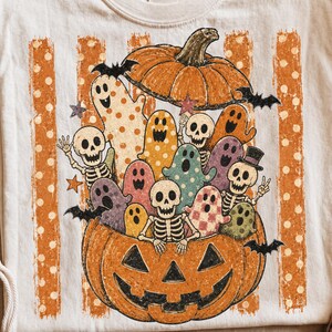May include: A cream-colored t-shirt featuring a graphic print of a large carved jack-o'-lantern filled with various colorful ghosts, skeletons, and bats. The design includes vertical orange polka-dot stripes and small star accents in a retro style.