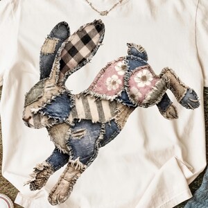 Op de afbeelding: Wit T-shirt met een patchwork konijn ontwerp. Het konijn is gemaakt van denim, ruitjes en bloemenstoffen in blauw, roze en bruin. Het shirt heeft een ronde hals en korte mouwen. De merknaam "Comfort Colors" staat op het label.