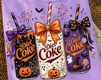 May include: A light purple t-shirt featuring a graphic of three Diet Coke cans decorated with Halloween themes. The cans are adorned with orange bows, straws, spider webs, pumpkins, ghosts, bats, and a witch hat, with fireworks in the background.