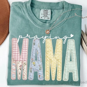 May include: A seafoam green t-shirt with the words "Praying MAMA" in white script and patchwork letters. The letters are in pink gingham, floral, light blue floral, yellow polka dot, and green gingham patterns. A silver necklace with a pendant rests on the shirt.