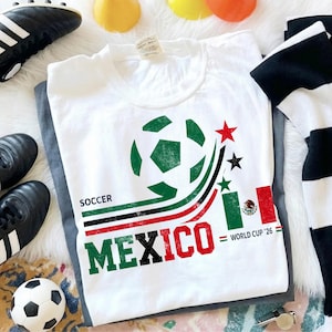 May include: A white t-shirt featuring a graphic of a soccer ball, curved green, black, and red lines, and the text "SOCCER MEXICO" in block letters. The design includes a small Mexican flag, three stars, and the text "WORLD CUP '26".