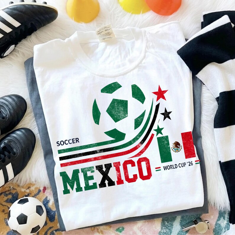 May include: A white t-shirt featuring a graphic of a soccer ball, curved green, black, and red lines, and the text "SOCCER MEXICO" in block letters. The design includes a small Mexican flag, three stars, and the text "WORLD CUP '26".