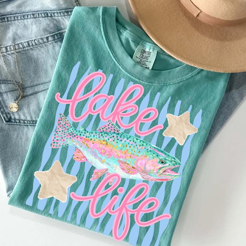 May include: Teal-colored t-shirt with the words "Lake Life" in pink script, a colorful fish graphic, and light blue wavy lines. Two tan star-shaped embellishments are also present. The shirt is folded on top of blue jeans and a tan hat.