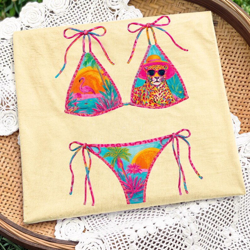 May include: A colorful bikini set with a tropical theme. The top features a flamingo and a leopard wearing sunglasses, while the bottom has a sunset and palm trees. The bikini is tied with pink and blue strings.