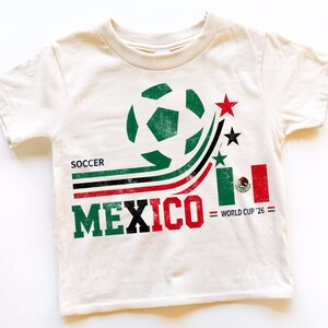 May include: A cream-colored short-sleeve t-shirt featuring a graphic print with a green soccer ball, curved stripes in green, black, and red, three stars, the text SOCCER MEXICO, a small Mexican flag, and the text WORLD CUP '26 in black and green.